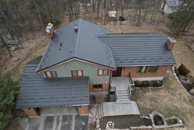 Drone angle with Hy-Grade metal roof in dark brown. The house in multi style, multiple stories and has green siding and brown brick accents.